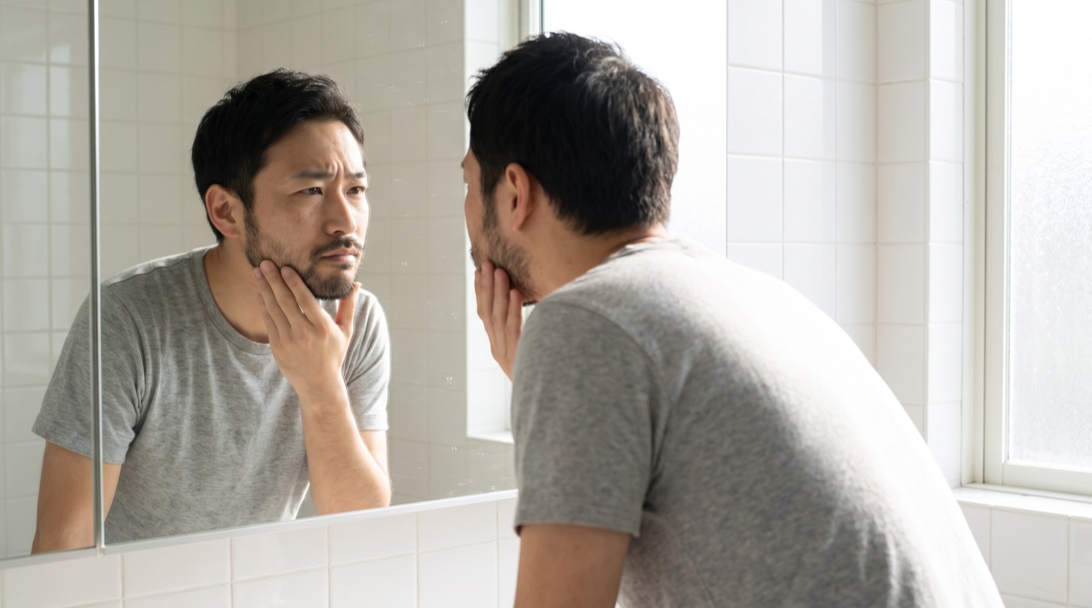 洗面所の鏡で自分の輪郭とあごのラインを手で確認する男性